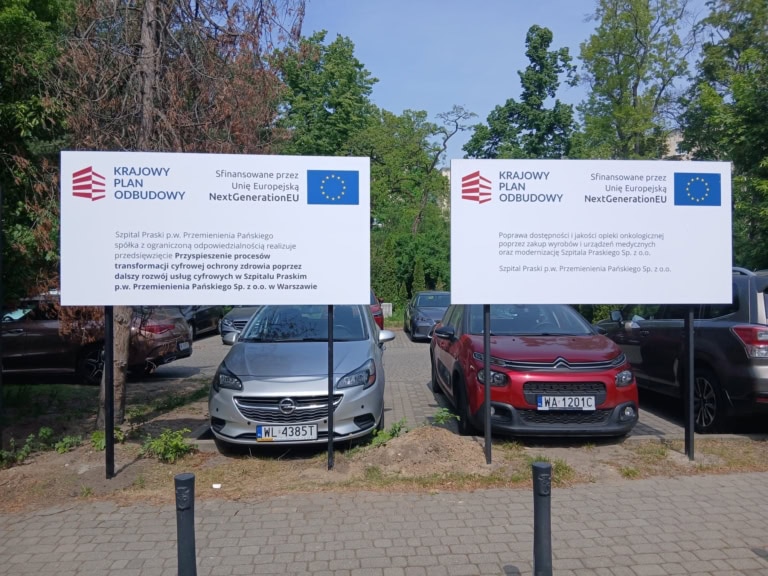 Trzy wolnostojące tablice informacyjne na czarnych słupach przed szpitalem